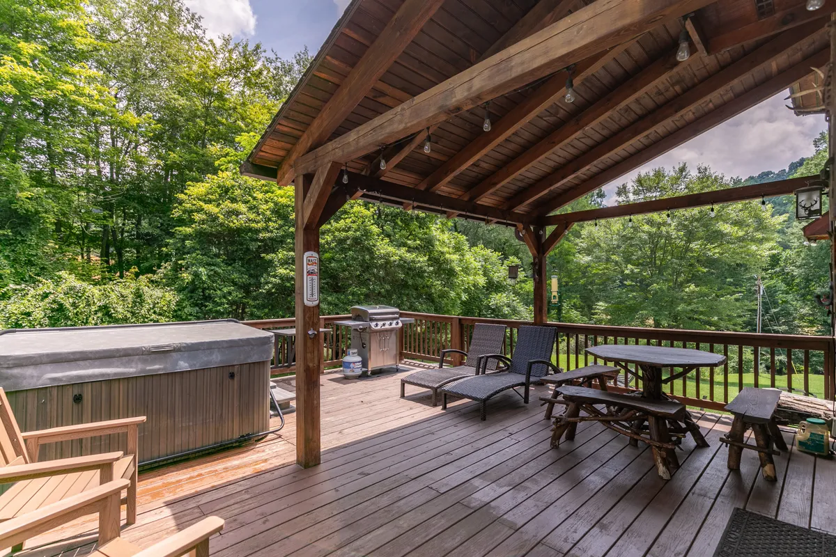 A bubbling outdoor hot tub sits on a wooden deck surrounded by lush greenery, with adjacent seating and a grill nearby. The scene suggests relaxation and socializing, with the promise of sunset views or starlit soaks in a serene mountain setting.
