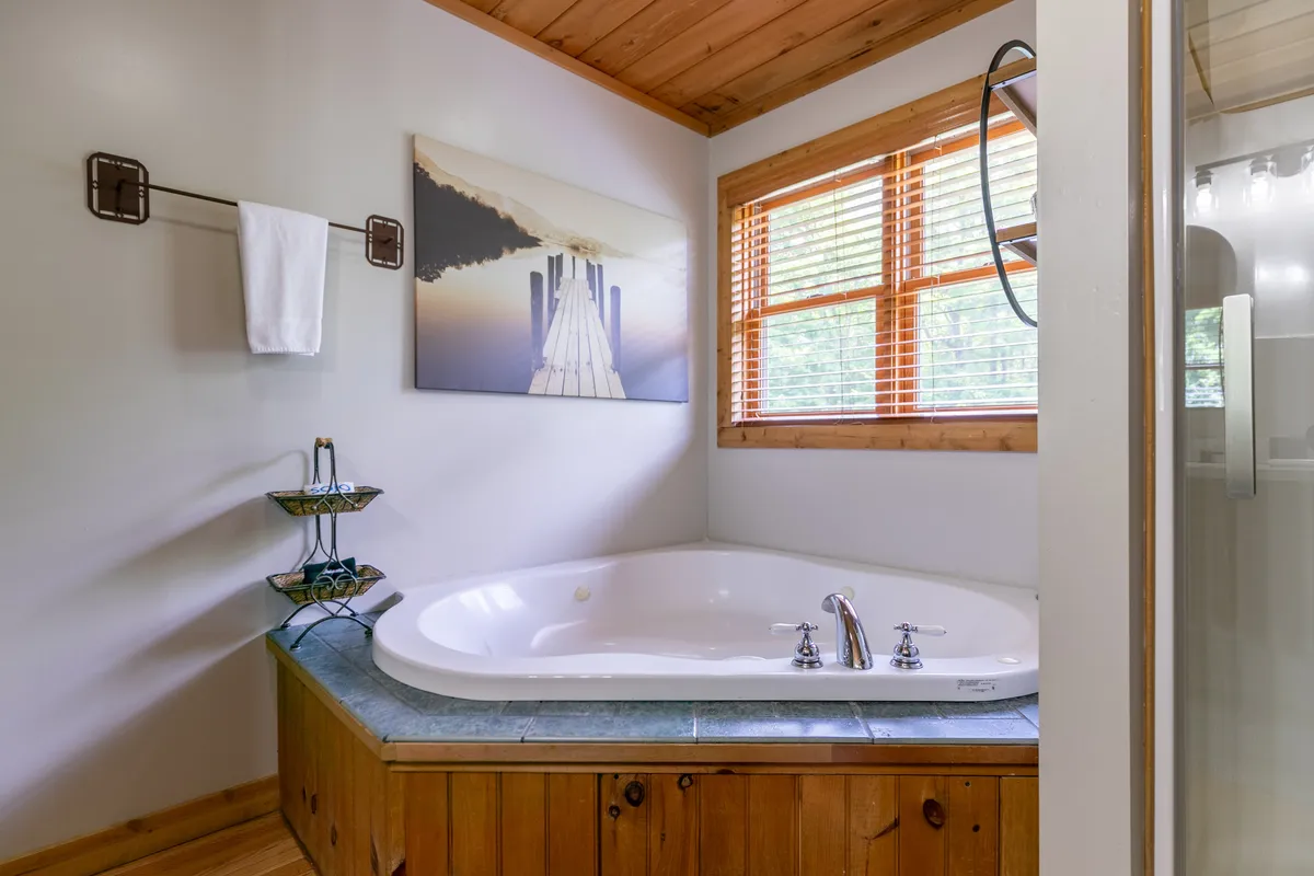The image features a whirlpool tub by a window with wooden blinds and a decorative painting. The atmosphere is calming and ideal for a spa-like escape, offering a glimpse into the upgraded king suite bathroom.