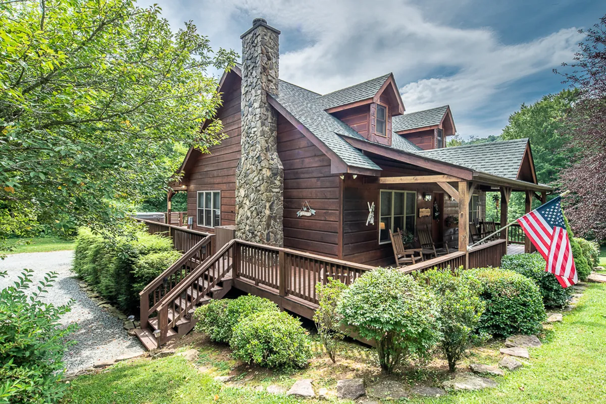 The image displays a welcoming wooden porch with lush greenery, a stone chimney, and an American flag. The cozy porch offers a spot for relaxation, framed by nature and perfect for unwinding after a day exploring the mountains.