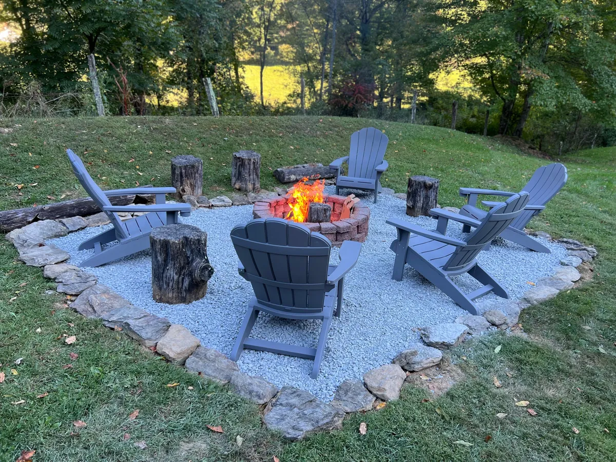 A classic wood-burning fire pit encircled by Adirondack chairs, natural stone borders, and stacked logs, set against a backdrop of trees and open yard. The setup invites groups to gather for storytelling, marshmallow roasting, or stargazing under the open sky.