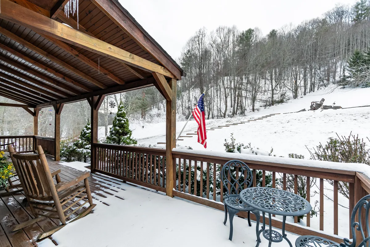 A pair of wooden rocking chairs and a small round table sit on a porch overlooking a snow-dusted mountain landscape and greenery. The American flag waves gently, and the scene feels tranquil and serene, perfect for peaceful moments outdoors.