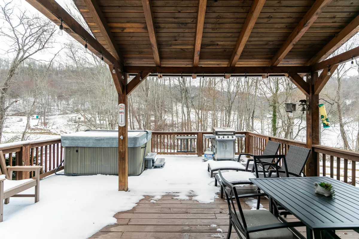 This exterior image showcases a spacious deck with a bubbling hot tub, a dining table set for meals, a grill, and sturdy wooden beams. The scene hints at both relaxation and outdoor adventure, with the deck inviting guests to enjoy the crisp mountain air and stunning natural vistas. In winter, snow blankets the scenery, enhancing the sense of a cozy retreat after outdoor activities.