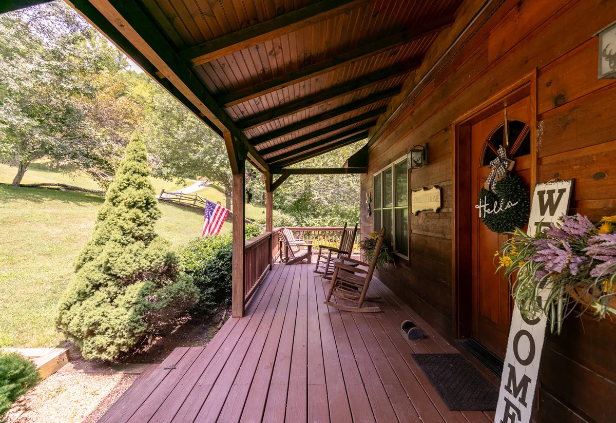 A welcoming, oversized covered front porch with classic rocking chairs, a visible welcome sign, an American flag, and touches of seasonal greenery and flowers. The porch exudes a cozy, inviting atmosphere, ideal for relaxing or social gatherings while enjoying the mountain air.
