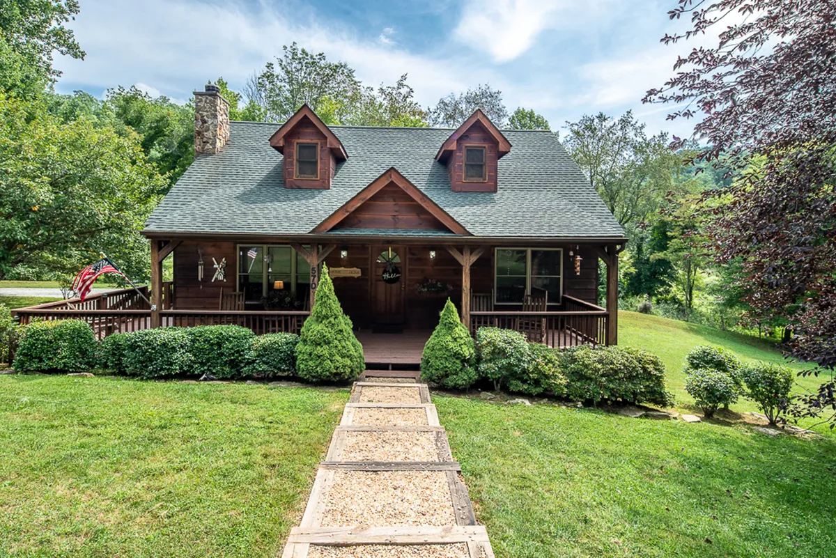 A wide view of the charming wooden house, complete with a stone chimney and manicured lawn, surrounded by mature trees. The open yard offers ample space for children and pets to play while establishing the privacy and peacefulness of the mountain setting.