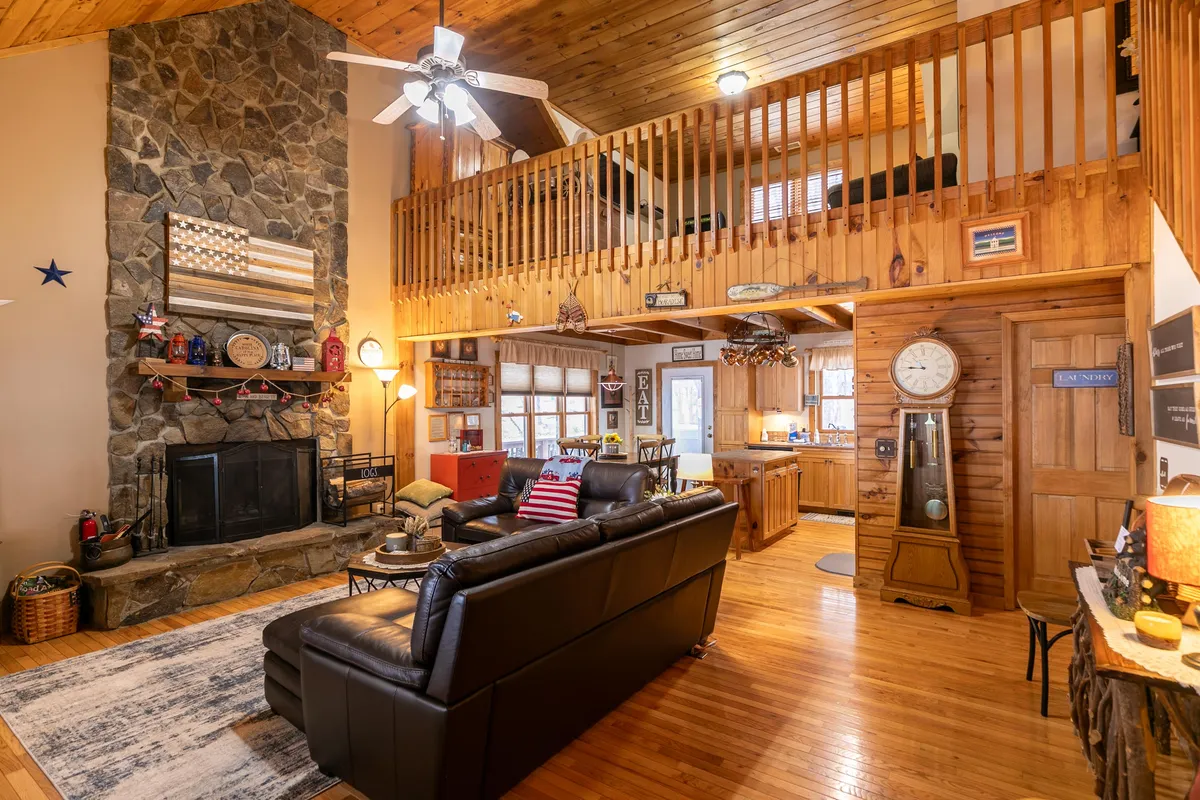 A warm, inviting living room with a prominent stone fireplace, soft leather sofa, and rustic wooden accents. The loft area above adds spaciousness, while decorative items and comfortable furnishings create a cozy, family-friendly setting ideal for gatherings and relaxation.