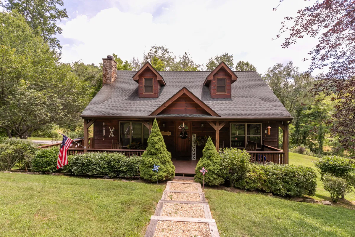 The image showcases the charming exterior of a cabin in Vilas, North Carolina, featuring a spacious front porch adorned with a welcome sign, a prominent stone chimney, manicured landscaping, and an American flag. The cozy ambiance invites guests to relax on the porch, enjoy the scenic outdoors, and experience the warmth of country living.
