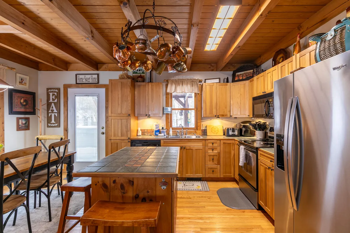 A cozy kitchen scene featuring a central island with a tiled countertop, wooden cabinetry, a dining table with chairs, and a hanging pot rack. The layout encourages conversation and meal preparation, inviting guests to gather and participate.