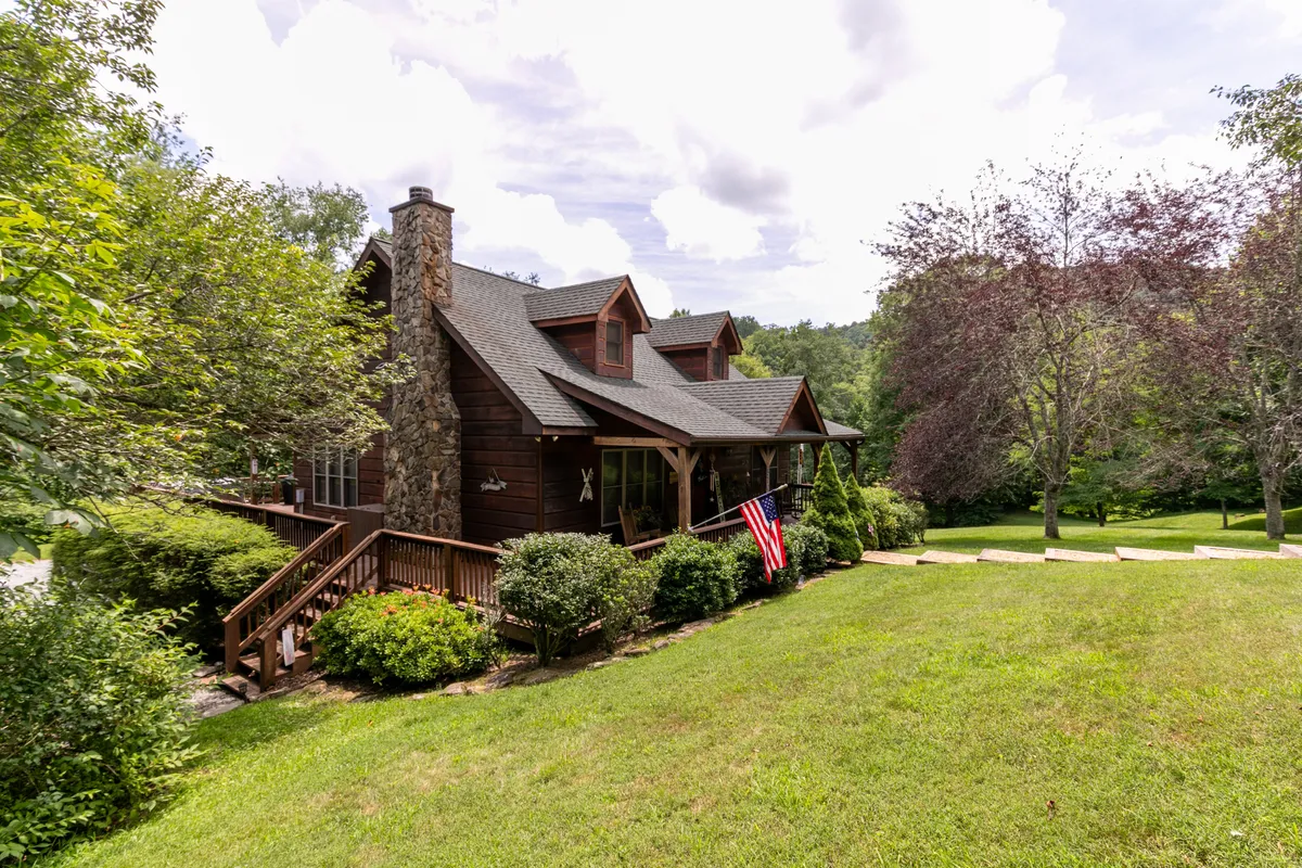 A wide view of the cabin's wooden front porch, accented by a classic stone chimney and an American flag. Lush greenery surrounds the property, creating a sense of seclusion and natural beauty. The porch offers comfortable seating, inviting relaxation and connection with the outdoors.