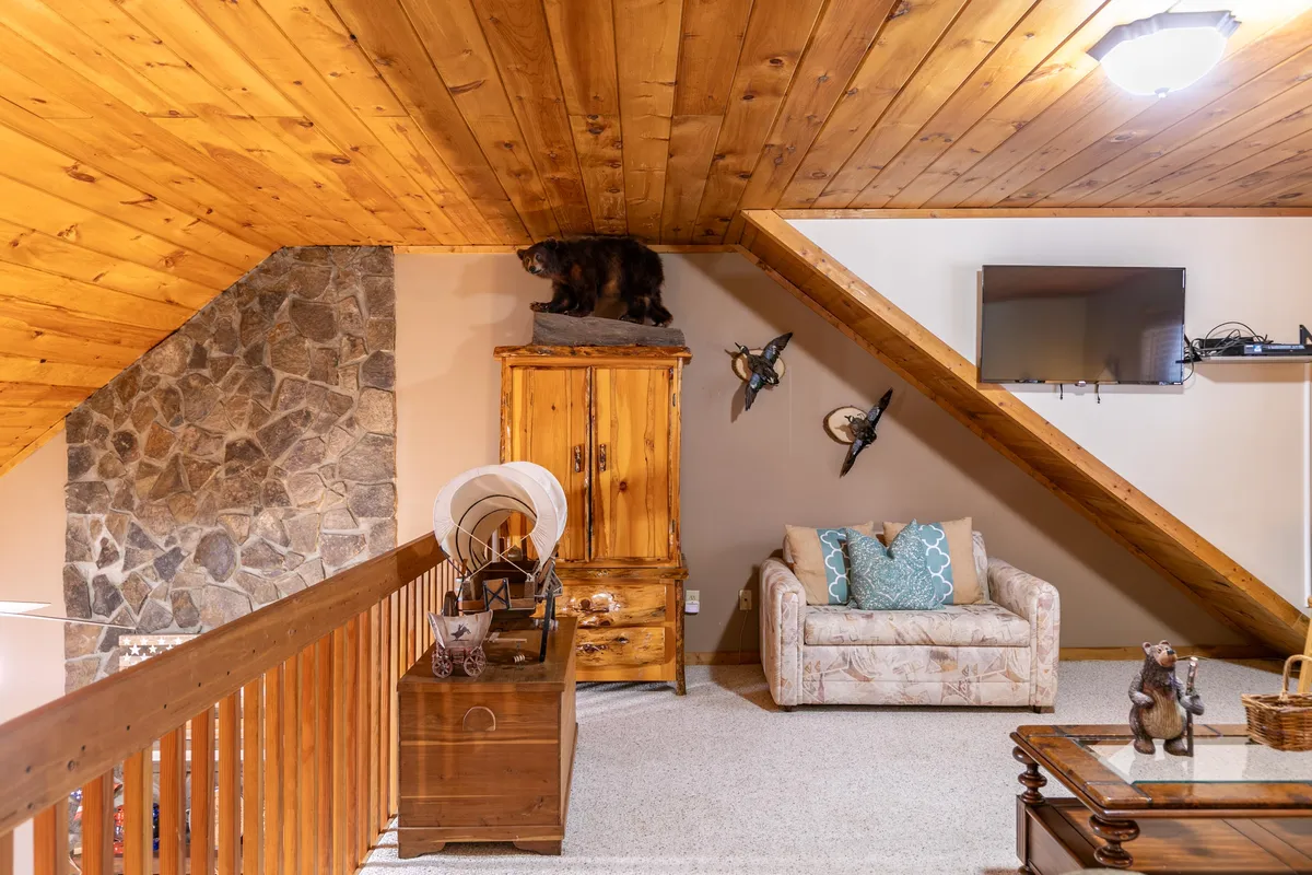 A cozy loft space with a wooden ceiling, bear mount, decorative furniture, and a comfortable couch with cushions. The inviting arrangement suggests a multipurpose area perfect for relaxing, reading, or serving as extra sleeping space for families.